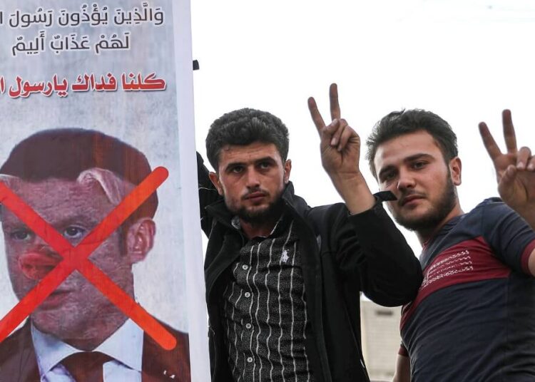 Men flash the victory gesture as they pose for a picture next to a sign condemning French President Emmanuel Macron, and depicting him with a pig snout, for his comments over Prophet Mohammed cartoons in Syria's rebel-held northwestern city of Idlib on October 25, 2020. (Photo by OMAR HAJ KADOUR / AFP)